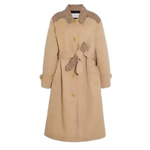 A long beige trench coat with button closure and a sash belt, featuring patterned accents on the shoulders and belt. It has cuffed sleeves and a classic collar design.