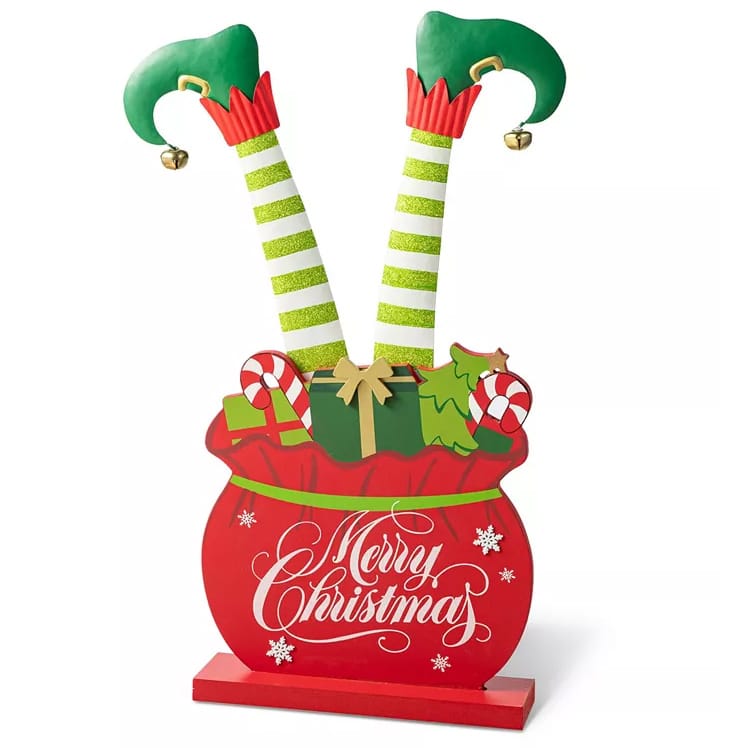 Festive porch decor featuring an elf's legs with curly shoes popping out of a red gift box adorned with candy canes and presents, and displaying \“Merry Christmas\“ text, enhanced with glitter and jingle bells.