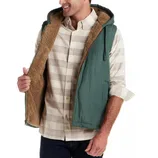 A man wears a green hooded vest with a brown fleece lining over a plaid button-up shirt, with dark jeans.