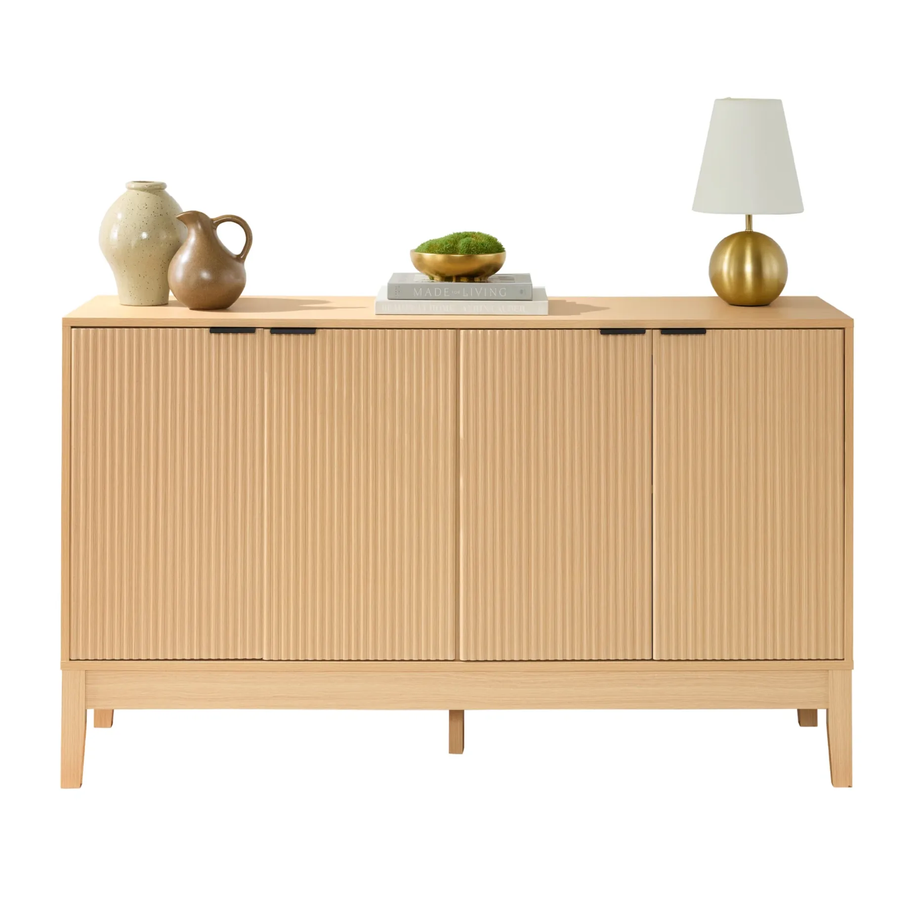 A light wood sideboard with vertical grooves features two pottery vases, two stacked books, a bowl with greenery, and a gold lamp on top.