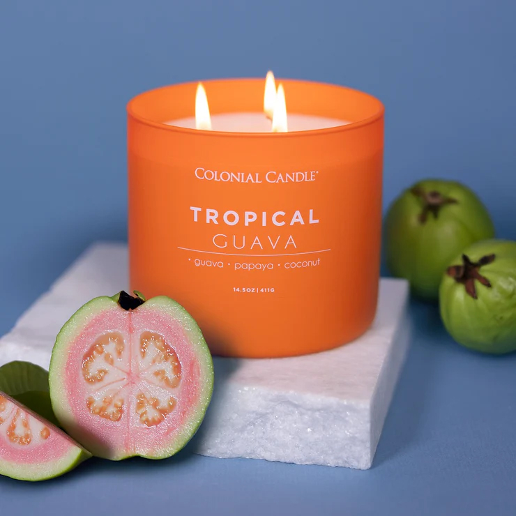 Orange-scented candle by Colonial Candle, labeled \“Tropical Guava,\“ featuring guava, papaya, and coconut, alongside fresh guavas on a white base.