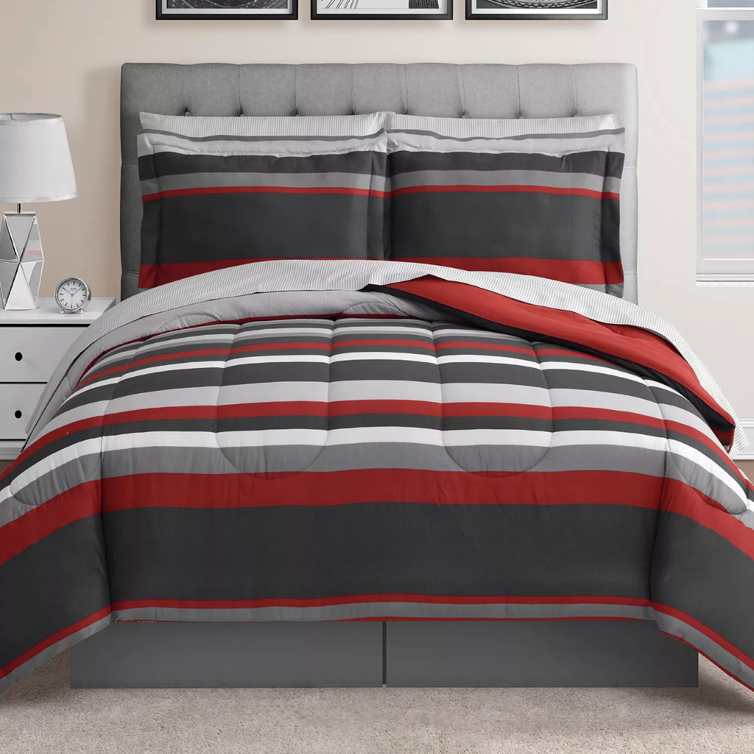 A bed is shown with a striped comforter set featuring red, gray, and white colors, accompanied by matching pillow shams. The bed is styled with a modern, tufted gray headboard, and a bedside table with a geometric lamp and clock.