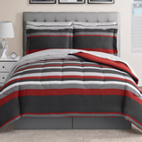 A bed is shown with a striped comforter set featuring red, gray, and white colors, accompanied by matching pillow shams. The bed is styled with a modern, tufted gray headboard, and a bedside table with a geometric lamp and clock.