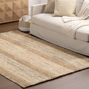 A textured beige and brown area rug lies on a wooden floor beside a light beige sofa with knitted throw pillows and a blanket.