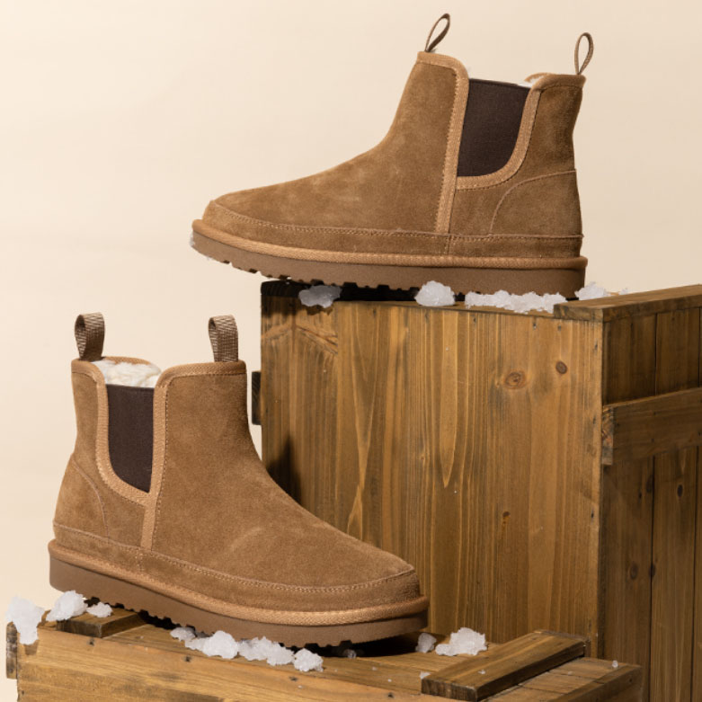 Brown suede Chelsea boots with white fur lining, featuring pull tabs and elastic side panels, displayed on wooden crates with small snow accents.
