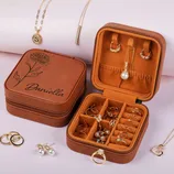 A brown leather jewelry case engraved with \“Daniella\“ contains rings, earrings, and necklaces, including pearl and gold pieces, displayed alongside additional jewelry items.