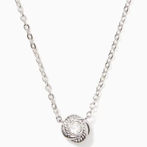 A silver chain necklace with a small, heart-shaped pendant that features an infinity symbol looped around a central, circular crystal detail.