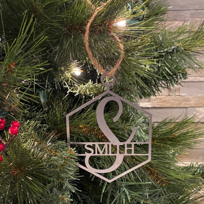 A holiday ornament with the initial \“S\“ and the name \“Smith,\“ made from a metallic material, hangs on a Christmas tree with evergreen branches and red berries.