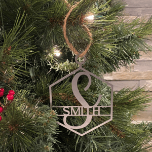 A holiday ornament with the initial \“S\“ and the name \“Smith,\“ made from a metallic material, hangs on a Christmas tree with evergreen branches and red berries.