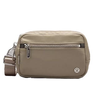 A beige crossbody bag with a front zipper pocket and an adjustable strap, featuring a circular logo on the bottom right.