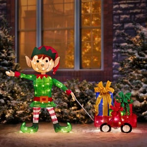 A festive light-up decoration features an elf in a green and red costume pulling a red wagon with two wrapped gifts, set against a snowy backdrop with trees and a lit house window.