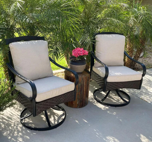 Two beige-cushioned swivel patio chairs with black metal frames and wicker bases are shown. They are placed outdoors on a concrete surface, accompanied by a small wooden table with a pink potted plant.