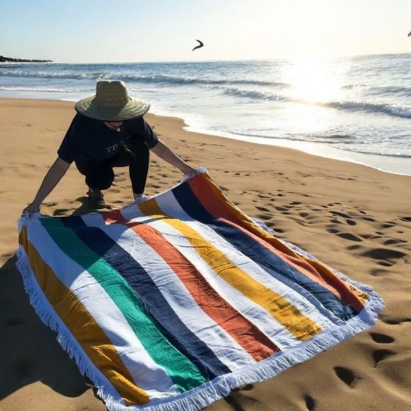 A beach towel featuring colorful vertical stripes in shades of green, blue, orange, and yellow, with white fringed edges, on sandy beach next to ocean. Dimensions: 60\“ x 60\“.