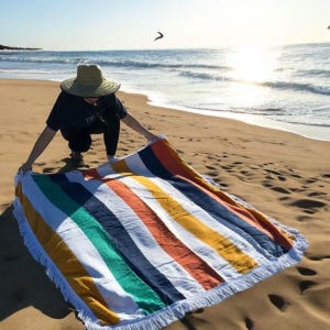 A beach towel featuring colorful vertical stripes in shades of green, blue, orange, and yellow, with white fringed edges, on sandy beach next to ocean. Dimensions: 60\“ x 60\“.