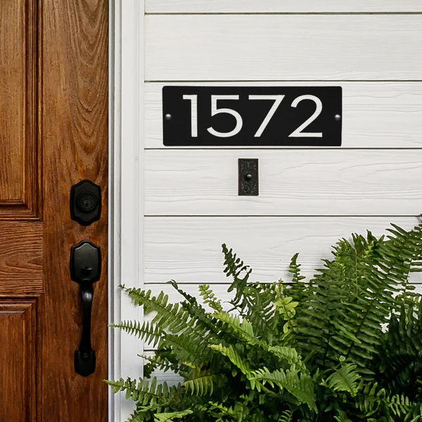 A black 9\“ horizontal address sign with white numbers \“1572\“ mounted on white siding next to a wooden door, accompanied by green plants below.