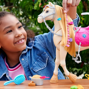 An interactive toy horse with a white coat and blonde mane, featuring accessories like a pink saddle and hair braiding tools. A child is decorating its mane with colorful clips. It includes grooming items like a brush, a bucket, and food accessories.