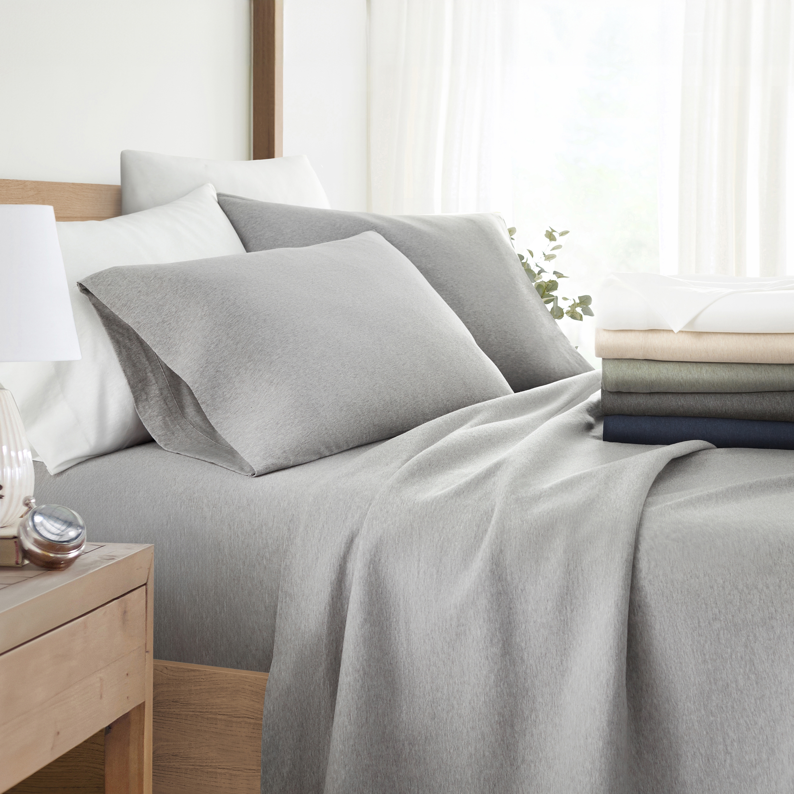 A neatly made bed with grey bedding, including sheets, pillowcases, and a comforter, beside a stack of folded linens in varying colors.