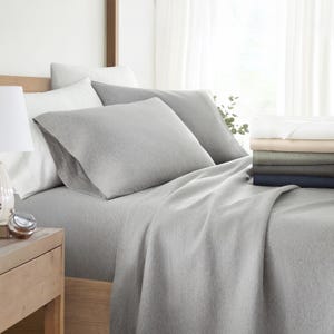 A neatly made bed with grey bedding, including sheets, pillowcases, and a comforter, beside a stack of folded linens in varying colors.