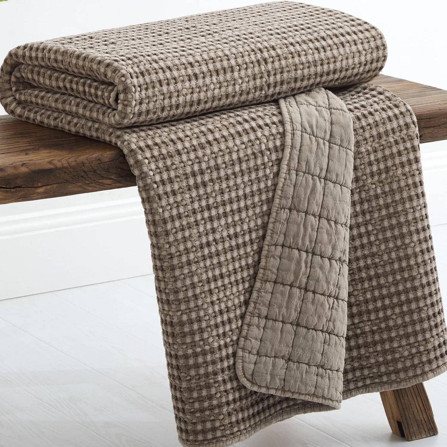 A folded taupe-colored textured blanket with a waffle pattern is draped over a wooden bench.