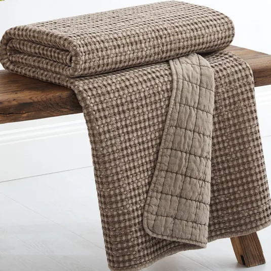 A folded taupe-colored textured blanket with a waffle pattern is draped over a wooden bench.