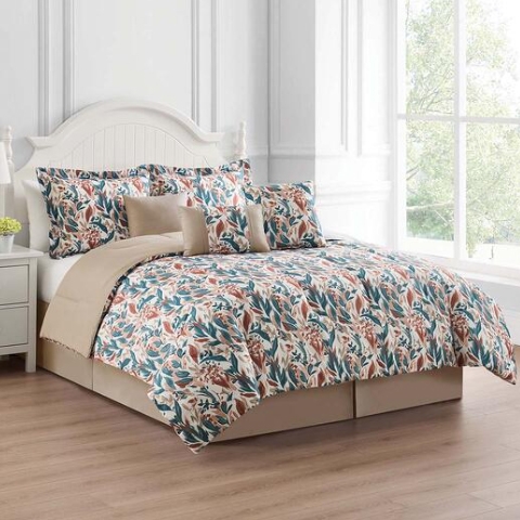 A floral-patterned bedding set with matching pillow shams and decorative pillows is displayed on a bed with a white headboard. The room has light wood flooring and a large window.