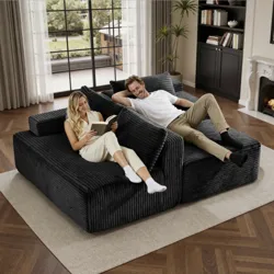 A cozy, large modular sofa with a textured black fabric is shown in a living room setting with two people relaxing. The space includes a bookshelf, a window with a view, and a fireplace.