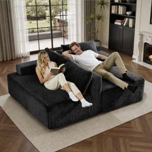 A cozy, large modular sofa with a textured black fabric is shown in a living room setting with two people relaxing. The space includes a bookshelf, a window with a view, and a fireplace.