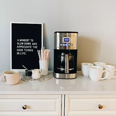 A Cuisinart 14-Cup Coffee Maker with a stainless steel carafe, a digital display, and backlit buttons, placed on a kitchen counter alongside mugs and a creamer container.