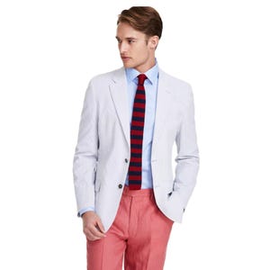 A man is wearing a light-colored blazer, blue dress shirt, red and navy striped tie, and pink trousers.