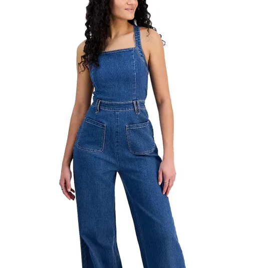 A woman is wearing a denim jumpsuit with wide legs, a square neckline, and large front pockets. The outfit is sleeveless with straps over the shoulders.