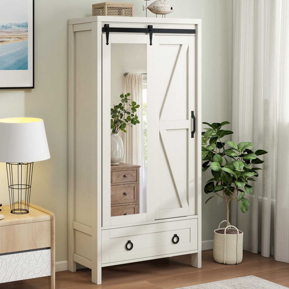 A white wardrobe with a sliding mirrored door features black hardware and a lower drawer, placed in a room with a table lamp, potted plant, and artwork.