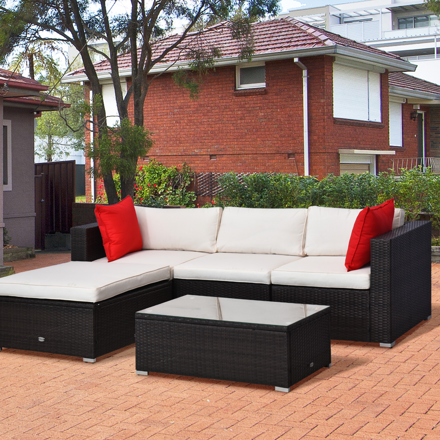 Outdoor patio set with a black wicker frame and cream cushions, featuring a sectional sofa, a matching chaise lounge, two red accent pillows, and a glass-top coffee table, set on a paved patio near a brick house and garden.