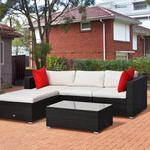 Outdoor patio set with a black wicker frame and cream cushions, featuring a sectional sofa, a matching chaise lounge, two red accent pillows, and a glass-top coffee table, set on a paved patio near a brick house and garden.