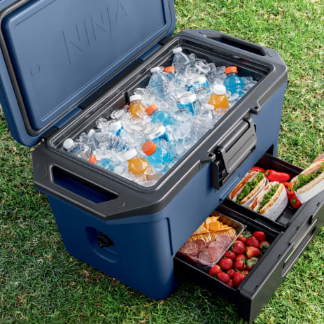 The Ninja 50gal FrostVault Cooler is shown open, with a compartment filled with ice, water, and sports drinks, alongside two drawers containing sandwiches, strawberries, and packaged snacks.