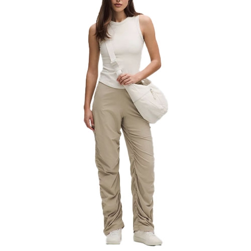 A woman is wearing a sleeveless white top and beige ruched pants, paired with white sneakers and carrying a matching beige crossbody bag.