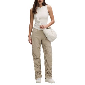 A woman is wearing a sleeveless white top and beige ruched pants, paired with white sneakers and carrying a matching beige crossbody bag.