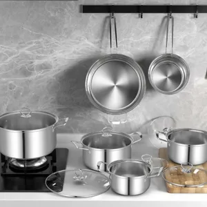 A set of stainless steel cookware is shown, including pots with lids on a stovetop, with two pans hanging on a wall. The setup is in a modern kitchen with a marble backsplash.