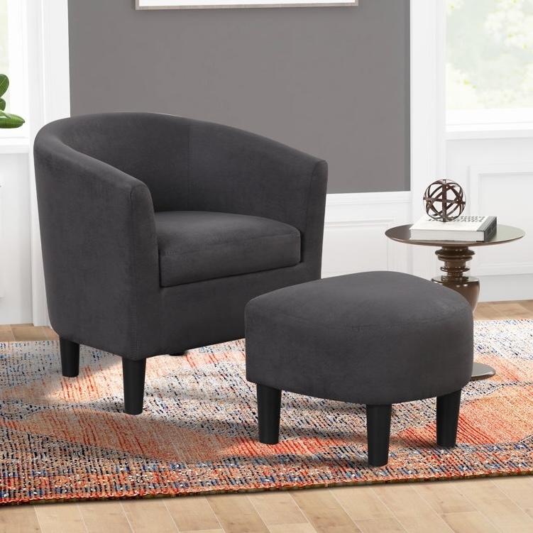 A dark gray upholstered tub chair with a matching ottoman is placed on a multicolored rug. Nearby is a round side table with decorative accents and books, all set against a gray wall and wooden floor.