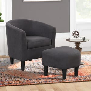 A dark gray upholstered tub chair with a matching ottoman is placed on a multicolored rug. Nearby is a round side table with decorative accents and books, all set against a gray wall and wooden floor.