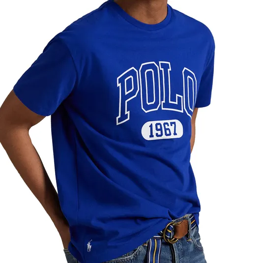 Royal blue t-shirt featuring \“POLO 1967\“ in large white lettering, paired with denim jeans and a striped belt. A small polo player logo is visible near the hem.