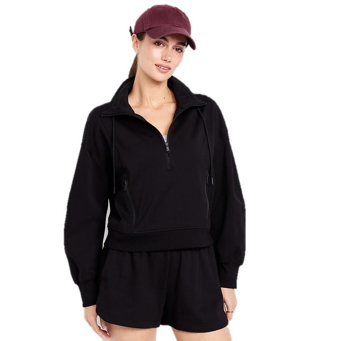 A woman is wearing a black zip-up pullover sweater with drawstrings and matching black shorts, paired with a maroon baseball cap.