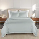 A bed setup with a light, striped duvet cover and two matching pillows, accompanied by modern nightstands with lamps, books, and decorative items on a soft, neutral-colored rug.