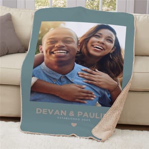 A cozy sherpa throw blanket featuring a photo of a smiling couple, with the text \“DEVAN & PAULINA ESTABLISHED 2025\“ printed below, along with a heart symbol. The blanket is blue with a soft beige lining on the reverse side and is displayed over a sofa.