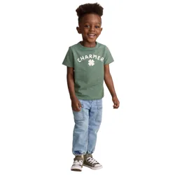 A young child is smiling, wearing a green \“CHARMER\“ t-shirt with a clover design, light blue jogger jeans, and olive green high-top sneakers with white laces.
