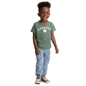 A young child is smiling, wearing a green \“CHARMER\“ t-shirt with a clover design, light blue jogger jeans, and olive green high-top sneakers with white laces.