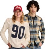 A person in a red cap and a cream sweater with \“90\“ stands next to another wearing a blue and yellow plaid shirt, both in casual attire.