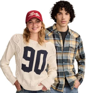 A person in a red cap and a cream sweater with \“90\“ stands next to another wearing a blue and yellow plaid shirt, both in casual attire.