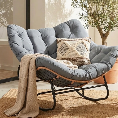 A cozy papasan chair with a gray cushion, featuring a decorative woven pillow and a beige blanket draped over it, placed on a woven rug near a potted plant.