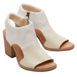TOMS Eliana Heeled Sandals feature a peep-toe design with a block heel, a neutral fabric upper, and side zippers for easy wear. The contrasting tan accents add a stylish touch, making them ideal for casual or semi-formal outfits.