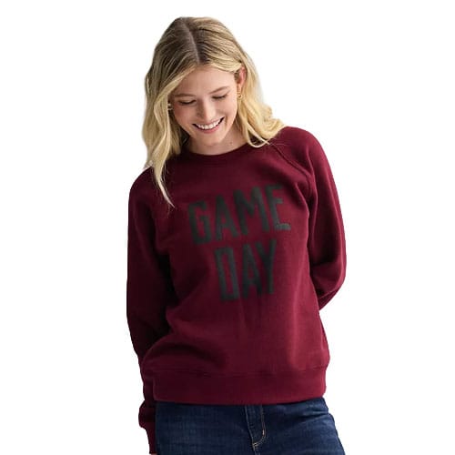 A woman is wearing a maroon sweatshirt with \“GAME DAY\“ printed on the front, paired with blue jeans.